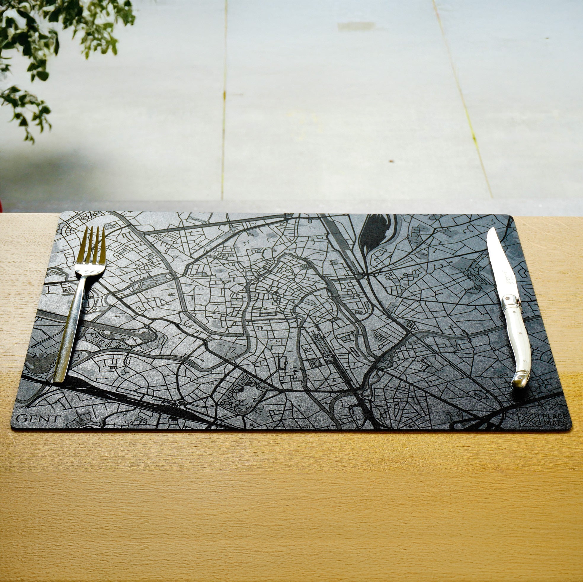 Een zwarte placemat van Place Maps met een gegraveerde plattegrond van Gent, klaargelegd met bestek op een houten tafel.