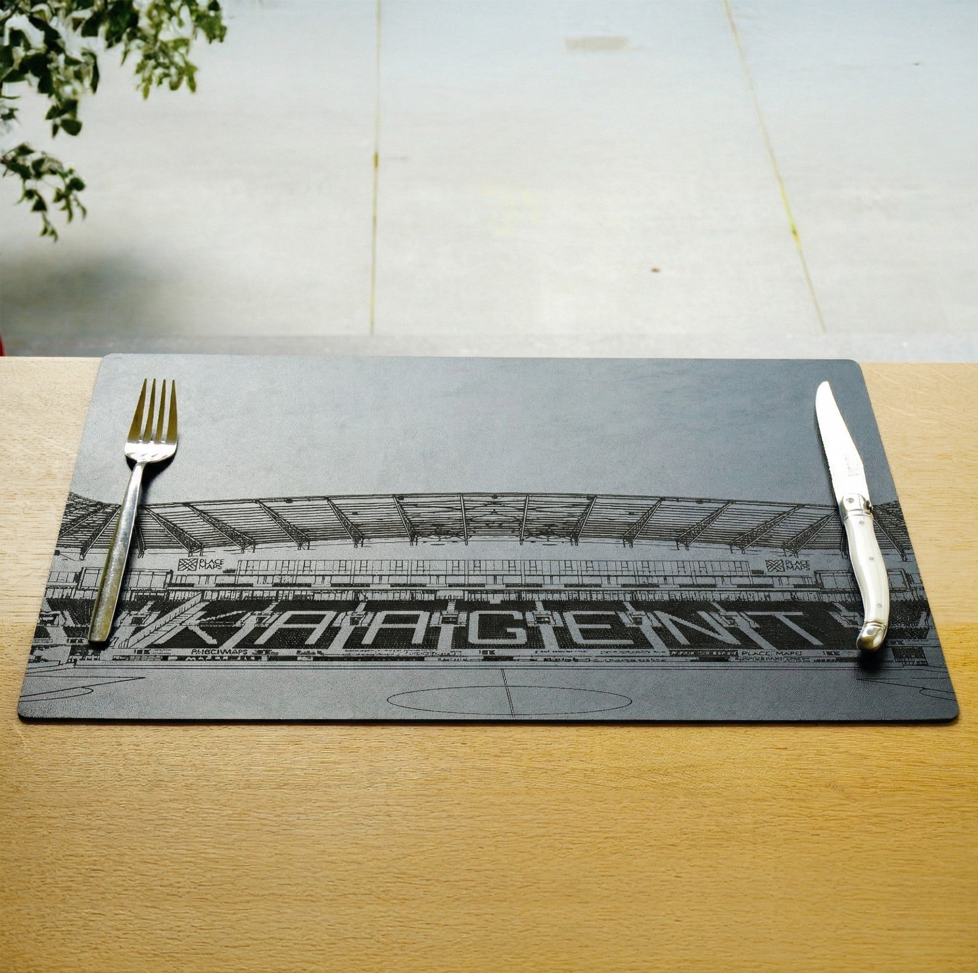 Een zwarte placemat van Place Maps met een gedetailleerde gravering van het KAA Gent stadion, klaargelegd met bestek op een houten tafel.